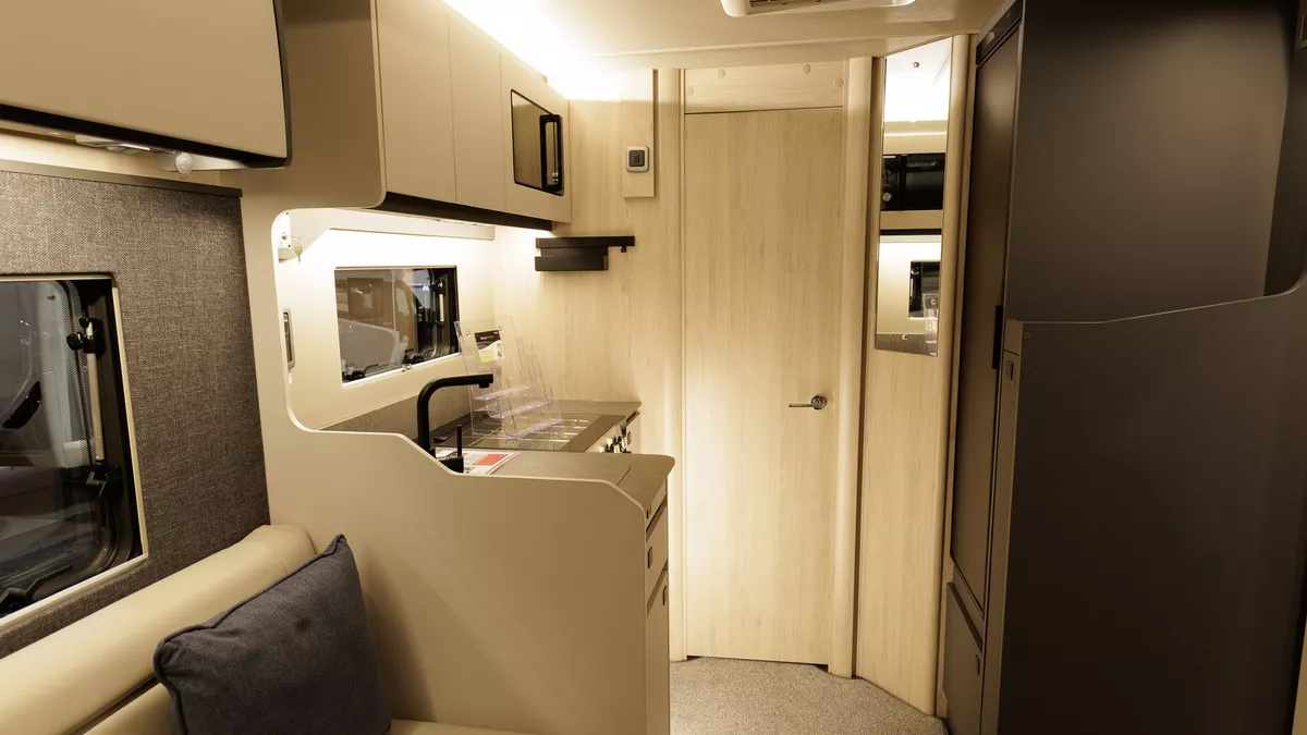 The Auto-Trail F-Line F67 has cream leather upholstery with pale wooden furniture and fittings.  The kitchen is on the left and there is an extractor fan on the ceiling.  The floor has a grey carpet.